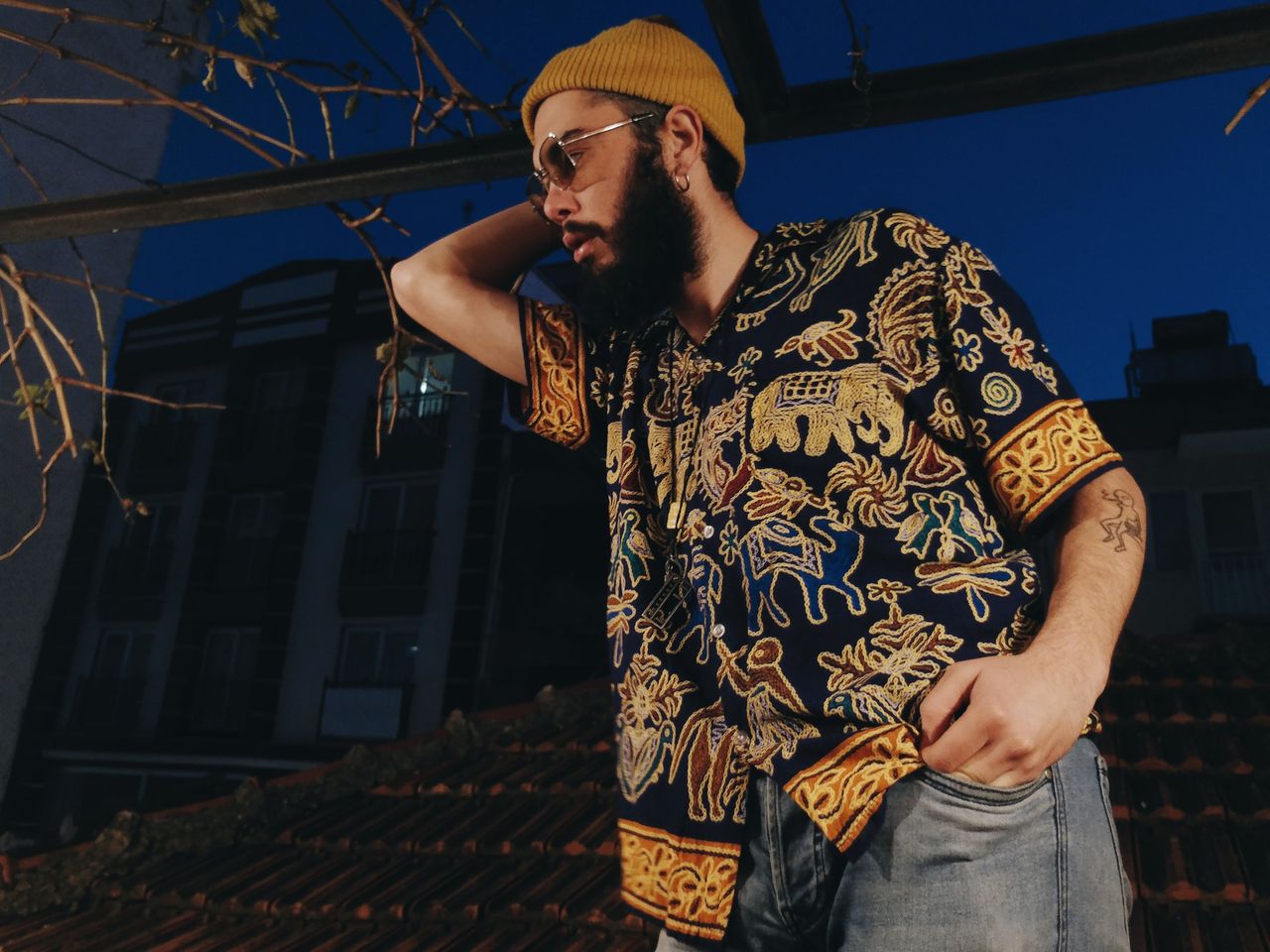 A fashionable man with a full beard, beanie, and sunglasses poses outdoors at night.